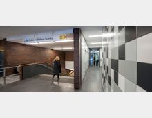 Untergrundbahnhof mit braunen Kachelwänden und einer Rolltreppe sowie einer Treppe, die nach unten führt. Rechts befindet sich ein Fahrstuhl mit einer Person und einem Kind davor, daneben eine Wand in schwarz-weißer Kacheloptik. Ein Schild oben zeigt die U7 in Richtung Rudow und Rathaus Spandau an.