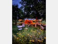 Ein Außenbereich mit mehreren Holztischen, Stühlen und Sonnensegeln, umgeben von Bäumen und Blumen. Im Hintergrund stehen eine rustikale Backsteinwand und warm leuchtende Lichterketten.