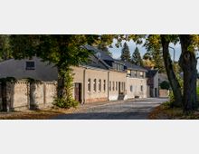 Eine malerische Kopfsteinpflasterstraße mit Wohnhäusern und einer alten Backsteinmauer auf der linken Seite, umgeben von Bäumen mit Herbstlaub. Im Hintergrund sind dichte Bäume und ein klarer, sonniger Himmel zu sehen.