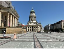 Weitläufiger Platz mit dem Deutschen Dom im Zentrum, einem barocken Gebäude mit Kuppel und Säulenfassade. Links steht das Konzerthaus Berlin mit Säulen und Statuen, umgeben von historischen Gebäuden und einer Kopfsteinpflasterfläche.