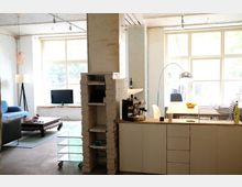 Helles Zimmer mit Loft-Charakter, aufgeteilt in einen Wohn- und Küchenbereich. Links ein Wohnbereich mit Sofa, Couchtisch und Fernseher, rechts eine offene Küche mit Arbeitsflächen, Regalen und großen Fenstern, die viel Tageslicht hineinlassen.