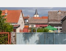 Gartenbereich mit Sicht auf mehrere Wohnhäuser mit roten und grauen Ziegeldächern. Im Hintergrund ist der Turm einer Kirche mit schwarzem Dach und goldener Spitze zu sehen.
