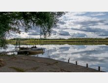 Ein ruhiger See mit spiegelglatter Wasseroberfläche, die den Himmel und Wolken reflektiert, umgeben von Wald am Ufer. Im Vordergrund befinden sich ein Steg mit einem anliegenden Motorboot und zwei Personen, die auf dem Steg sitzen.