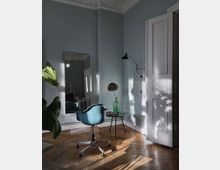 Ein heller Raum mit hellblauen Wänden, einem hölzernen Fischgrätboden und einer hohen weißen Zimmertür. Im Vordergrund steht ein blauer Drehstuhl vor einem Spiegel, daneben ein kleiner Tisch mit einer grünen Glasvase. Schatten von Pflanzen und Licht werden an die Wand projiziert.