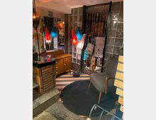 Ein gemütlicher, retro-inspirierter Innenraum mit gemustertem Sessel, schwarzem rundem Teppich, gläserner Ziegelwand und bunter Perlenvorhang-Abtrennung. Lampen mit bunten Schirmen und eine Holzkommode ergänzen den Raum, der mit Fliesenboden gestaltet ist.