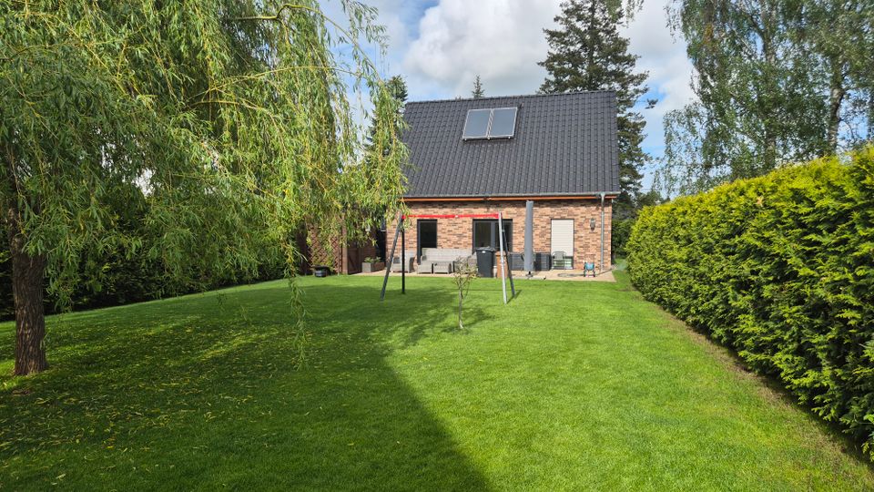 Rasenfläche in einem gepflegten Garten hinter einem Einfamilienhaus aus Klinker mit steilem, dunklem Satteldach und Dachfenstern. Der Garten ist von hohen Hecken und Bäumen eingerahmt und hat eine kleine Terrasse sowie ein schlichtes Schaukelgestell.