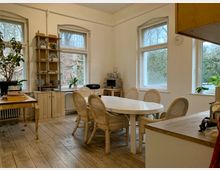 Heller Essbereich mit weißen Wänden, alten Holzdielen und großen Sprossenfenstern, die den Blick auf eine bewaldete Außenlandschaft freigeben. Mittig steht ein ovaler Esstisch mit geflochtenen Stühlen, umgeben von Regalen, Pflanzen und einer Küchenzeile im Hintergrund.