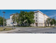 Wohnblock im Stil des sozialistischen Klassizismus an einer Straßenkreuzung mit Kopfsteinpflaster; im Vordergrund sind Laternen, Straßenschilder und Bäume zu sehen, die den Gebäudebereich säumen.