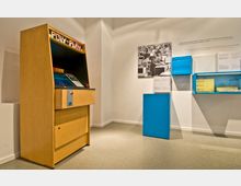 Ausstellungsraum mit einem Retro-Spielautomaten namens „Poly-Play“ aus Holz auf der linken Seite, einer Fotografie und Informationstafeln an der weißen Wand, sowie blauen Ausstellungsvitrinen und Podesten auf der rechten Seite.