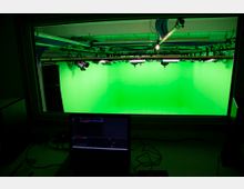 Ein Aufnahmestudio mit einem großen Green Screen, sichtbar durch ein Fenster von einem Kontrollraum aus. Im Vordergrund steht ein Laptop auf einem Schreibtisch, während Beleuchtungsausrüstung an der Studiodecke installiert ist.