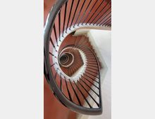 Spiralförmiges Treppenhaus mit roten Treppenstufen, weißen Wangen und einem schwarzen Geländer, das sich zentrisch nach unten windet. Die Perspektive zeigt einen Blick von oben in die Tiefe der Spirale.
