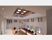 Großer, moderner Innenhof mit weißen Wänden und umlaufender Galerieebene, umgeben von zahlreichen Fenstern und Glasfronten. In der Decke befinden sich mehrere runde Oberlichter; unten ist ein Wartebereich mit Sitzbänken und digitalen Anzeigen.