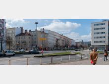 Straßenszene in einer Stadt mit mehrstöckigen Wohnhäusern im Altbau- und Neubau-Stil. Im Vordergrund verläuft eine belebte Kreuzung mit Autos, Fußgängern und einem gelben Straßenschild mit der Aufschrift „Charlottenburg“.