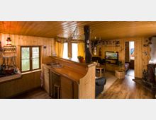 Gemütlicher Innenraum einer Holzhütte mit einer offenen Küche, Barbereich und Wohnzimmer. Die Einrichtung besteht aus Holzmöbeln, einem kleinen Kamin, gelben Vorhängen und Dekorationen wie Hängenetzen und Regalen.