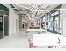 Heller, moderner Showroom mit weißen Wänden und großen Glasfenstern, in dem Kleidung an Kleiderständern präsentiert wird. Im Vordergrund befinden sich Tische und Stühle, die unter linearen Deckenleuchten stehen, während die Rückwand aus Sichtbeton gestaltet ist.