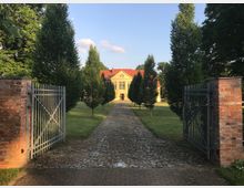 Gepflasterter Weg durch ein offenes Metalltor mit roten Ziegelpfeilern, flankiert von symmetrisch angeordneten Bäumen, führt zu einem beige-gelben Gebäude mit rotem Satteldach inmitten einer gepflegten Rasenfläche. Der Himmel ist klar mit ein paar Wolken sichtbar.