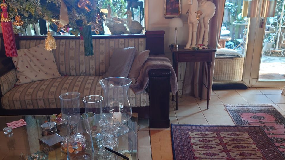 Wohnzimmer mit gemütlicher Einrichtung, bestehend aus einem Sofa mit gestreiften Kissen, einem Glastisch mit dekorativen Objekten und einer Weihnachtsdekoration mit Tannenzweigen. Im Hintergrund sind große Fenster und Glastüren zu einem Außenbereich mit Pflanzen sichtbar, daneben hängen Bilder und Vorhänge an den Wänden.