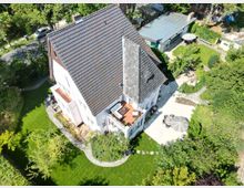 Luftaufnahme eines Einfamilienhauses mit weißer Fassade, steilem Ziegeldach und einem gepflegten Garten. Der Garten umfasst eine Terrasse mit Sonnenschirmen, Rasenflächen und mehreren Gehwegen, umgeben von Bäumen und Büschen.