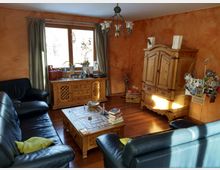 Wohnzimmer mit orangefarbenen Wänden, dunklen Ledersofas und Holzmöbeln, darunter ein Schrank, ein Couchtisch und eine Kommode unter einem Fenster mit Vorhängen. Die Einrichtung enthält persönliche Gegenstände, Bücher und Fotos, während Sonnenlicht durch das Fenster fällt.