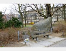 Eine Parkszene mit einer Holzbank vor einem großen Felsblock, umgeben von kahlen Bäumen und Sträuchern. Im Vordergrund steht ein Metallmülleimer, im Hintergrund sind ein Gebäude sowie weitere Grünflächen sichtbar.
