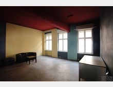 Minimalistisch eingerichteter Raum mit Betonboden, einer gelben und blauen Wand, sowie drei großen Fenstern mit weißen Rahmen. Im Vordergrund befinden sich ein braunes Ledersofa mit einem kleinen Tisch und rechts ein weißer Schreibtisch.