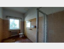 Ein Badezimmer mit einer veralteten Einrichtung, beige-orangefarbenen Fliesen an den Wänden und einem roten Fliesenboden. Es gibt ein Fenster mit Heizkörper darunter, ein WC, einen Spiegel, eine an der Wand montierte Ablage sowie eine Dusche mit gläserner Tür auf der rechten Seite.