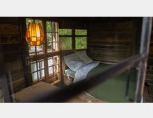 Rustikaler Innenraum einer Holzhütte mit einem Bett, das mit weißer Bettwäsche und einer grünen Decke ausgestattet ist. Große Fenster mit Holzrahmen lassen Tageslicht herein, und eine warme, orangefarbene Hängelampe beleuchtet den Raum dezent.