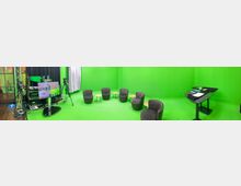 Ein Raum mit grüner Wand und grünem Boden, ausgestattet als moderner Greenscreen-Studio-Bereich. Im Vordergrund befinden sich mehrere gepolsterte Stühle und kleine runde Holztische, links sind Kameras, Monitore und technische Ausrüstung sichtbar.