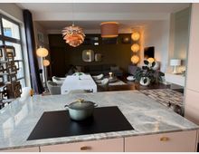 Open-plan modern kitchen and living area with a marble-topped island in the foreground, an induction cooktop, and a dining table leading toward a lounge with a sectional sofa. Large windows, multiple pendant and floor lamps, indoor plants, and a mix of rugs and wood flooring define the space.