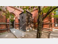 Backsteinfassade eines historischen Gebäudes mit verzierten Fensterbögen und moderner, kubisch geformter Anbau-Verkleidung aus Metall; im Vordergrund Bäume und ein befestigter Weg mit Rampe.