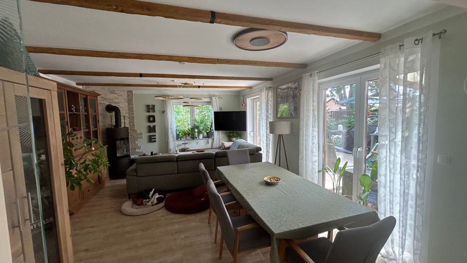 Offener Wohn- und Essbereich mit Holzbalkendecke, Esstisch mit Stühlen und angrenzender Sitzecke vor einem Kaminofen. Große bodentiefe Fenster mit hellen Vorhängen lassen viel Tageslicht herein und bieten Blick in den Garten; seitlich stehen Schränke und mehrere Zimmerpflanzen.