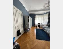 Ein geräumiges Zimmer mit einem Fischgrätenparkettboden, blauen Wänden und einer verzierten Decke mit Stuck. Der Raum enthält einen Kleiderschrank, ein Bett, einen Schreibtisch vor einem großen Fenster mit weißen Vorhängen und einen Kronleuchter an der Decke.