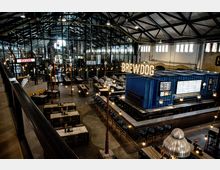 Großer, industriell gestalteter Innenraum einer Brauerei mit hohen Decken, freiliegenden Stahlträgern und Glasfassade. Im Vordergrund befinden sich Sitzbereiche mit Tischen, während im Hintergrund eine Bar aus blauen Schiffscontainern mit leuchtender "BrewDog"-Beschriftung und Zapfhähnen zu sehen ist.