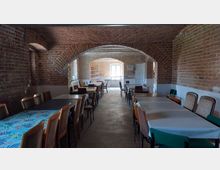 Ein länglicher Raum mit freiliegenden Ziegelwänden und gemauerten Gewölbedecken. Der Raum ist mit mehreren Tischen und Stühlen möbliert, und durch ein Fenster im Hintergrund fällt Tageslicht ins Innere.