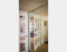 Ein Innenraum mit einer weißen Tür mit Glaseinsätzen auf der linken Seite und einer spiegelnden, geometrisch unterteilten Schrankwand auf der rechten Seite. Im Hintergrund ist ein heller Raum mit Regalen, Büchern und einem Tisch mit Stühlen zu sehen.