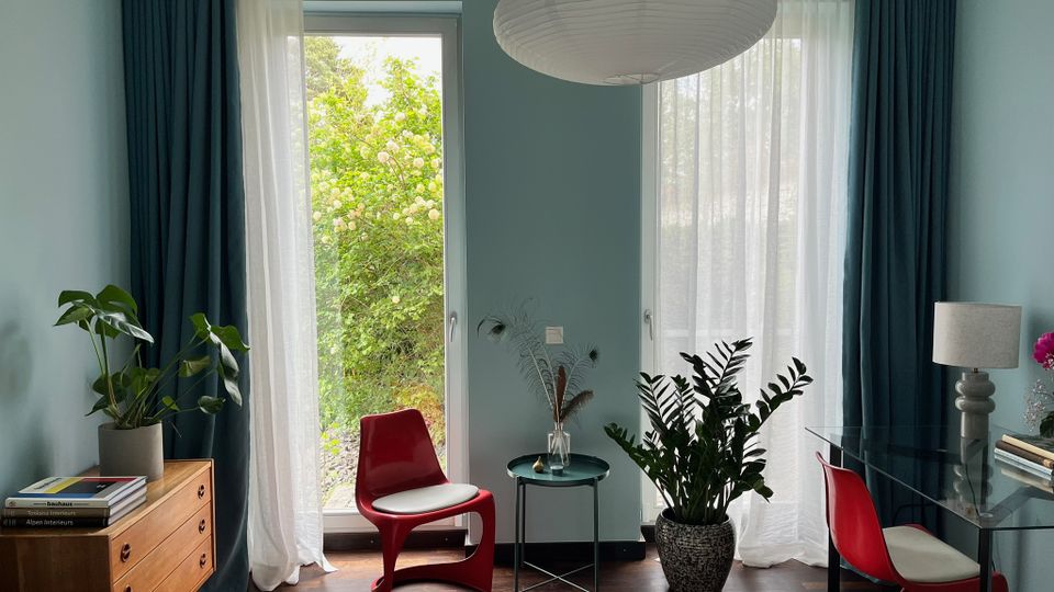 Modernes Schlafzimmer mit hellblauen Wänden, zwei bodentiefen Fenstern mit weißen Gardinen und dunklen Vorhängen sowie Blick ins Grüne. Im Raum stehen rote Stühle, ein Glastisch mit Lampe, mehrere Zimmerpflanzen, eine Holzkommode und eine große runde Papier-Deckenleuchte.