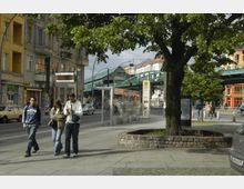 Städtische Straßenecke mit Bäumen und einer Bushaltestelle im Vordergrund. Im Hintergrund sind mehrstöckige Gebäude, ein erhöht verlaufendes Bahngleis sowie Passanten und parkende Autos zu sehen.