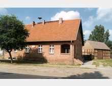 Ein einstöckiges Backsteinhaus mit rotem Satteldach, seitlich steht ein einfacher Holzschuppen im Fachwerkstil. Im Vordergrund befindet sich ein gepflasterter Gehweg, daneben ein Baum, der teilweise Schatten auf die Hauswand wirft.
