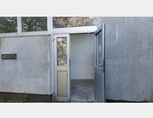 Eine geöffnete Tür eines schlichten Gebäudes mit grauer Putzfassade. Durch die Glaseinsätze der Tür und das Fenster darüber sind Bäume im Hintergrund erkennbar, der Boden vor dem Eingang besteht aus Beton.