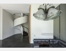 Innenraum mit minimalistischer Gestaltung, einer weißen, spiralförmigen Treppe vor einer schlichten Wand und einer Hängelampe mit großen, industriellen Strahlern. Rechts im Bild befindet sich eine schwarze Ledercouch vor einer weißen Wand.