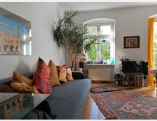 Wohnzimmer mit weiß gestrichenen Wänden, einem grauen Sofa mit bunten Kissen, einem großen Teppich mit geometrischen Mustern und einem großen Fenster mit Blick ins Grüne. Im Raum befinden sich Pflanzen, ein kleiner Tisch, eine Stereoanlage und verschiedene dekorative Elemente.