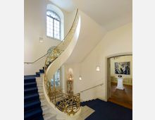 Eleganter Treppenaufgang in einem historischen Gebäude mit einer geschwungenen Treppe, goldverziertem Geländer und blauen Teppichläufern; im Hintergrund ein Raum mit Holzfußboden, einem Stehtisch und einem Wandgemälde.