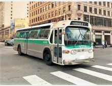 Ein Retro-Stadtbus mit grün-weißer Lackierung fährt durch eine Straßenkreuzung in einer städtischen Umgebung mit historischen Gebäuden im Hintergrund. Ein Zebrastreifen ist im Vordergrund zu sehen.
