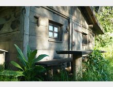 Holzhaus mit einer wettergegerbten Fassade, kleinen Fenstern mit hölzernem Rahmen und einer Holztür; davor eine einfache Bank und ein Tisch aus massivem Holz, umgeben von hohem Gras und üppigen Pflanzen. Im Hintergrund sind Bäume und Sträucher zu erkennen.