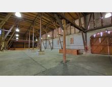 Großer Innenraum einer Scheune mit Holzbalkenkonstruktion, hellem Mauerwerk und einem Dach aus Holz. Der Raum ist nahezu leer, mit vereinzelten Holz- und Backsteinpodesten sowie einer großen Holztür auf der rechten Seite.