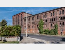 Backstein-Industriegebäude mit mehreren Stockwerken und schmalen Rundbogenfenstern, teilweise von Efeu bewachsen. Davor eine Straße und ein Bürgersteig mit Straßenlaterne sowie eine Hecke auf der linken Seite.