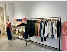 Ein heller Ladenraum mit Kleiderständer, der farblich sortierte Jacken, Pullover und T-Shirts präsentiert. Rechts steht eine Schaufensterpuppe mit einem rosa Oberteil, während links Regale mit gefalteter Kleidung und hängenden Körben zu sehen sind.