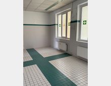 Leerer, gekachelter Raum mit weißen Wandfliesen und einem grünen Fliesenband sowie grün-weiß gemustertem Fliesenboden. Zwei Fenster mit Milchglas und Erste-Hilfe-Kreuz-Symbolen sowie darunterliegende Heizkörper prägen die Seite des Raums.