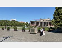 Ein öffentlicher Platz mit Pflastersteinen, im Hintergrund das Alte Museum in Berlin mit seinen klassischen Säulen und der Rasenfläche davor. Links ist ein Brunnen sichtbar, umgeben von Bäumen, mit vereinzelten Menschen im Bereich.