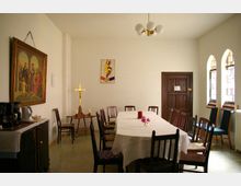 Ein schlicht eingerichteter Gemeinderaum mit einem langen, weiß gedeckten Tisch, umgeben von Holzstühlen. Linke Wand mit religiösem Gemälde und Kreuz, rechte Seite mit drei bogenförmigen Fenstern aus Buntglas, sowie einer Holztür im Hintergrund.